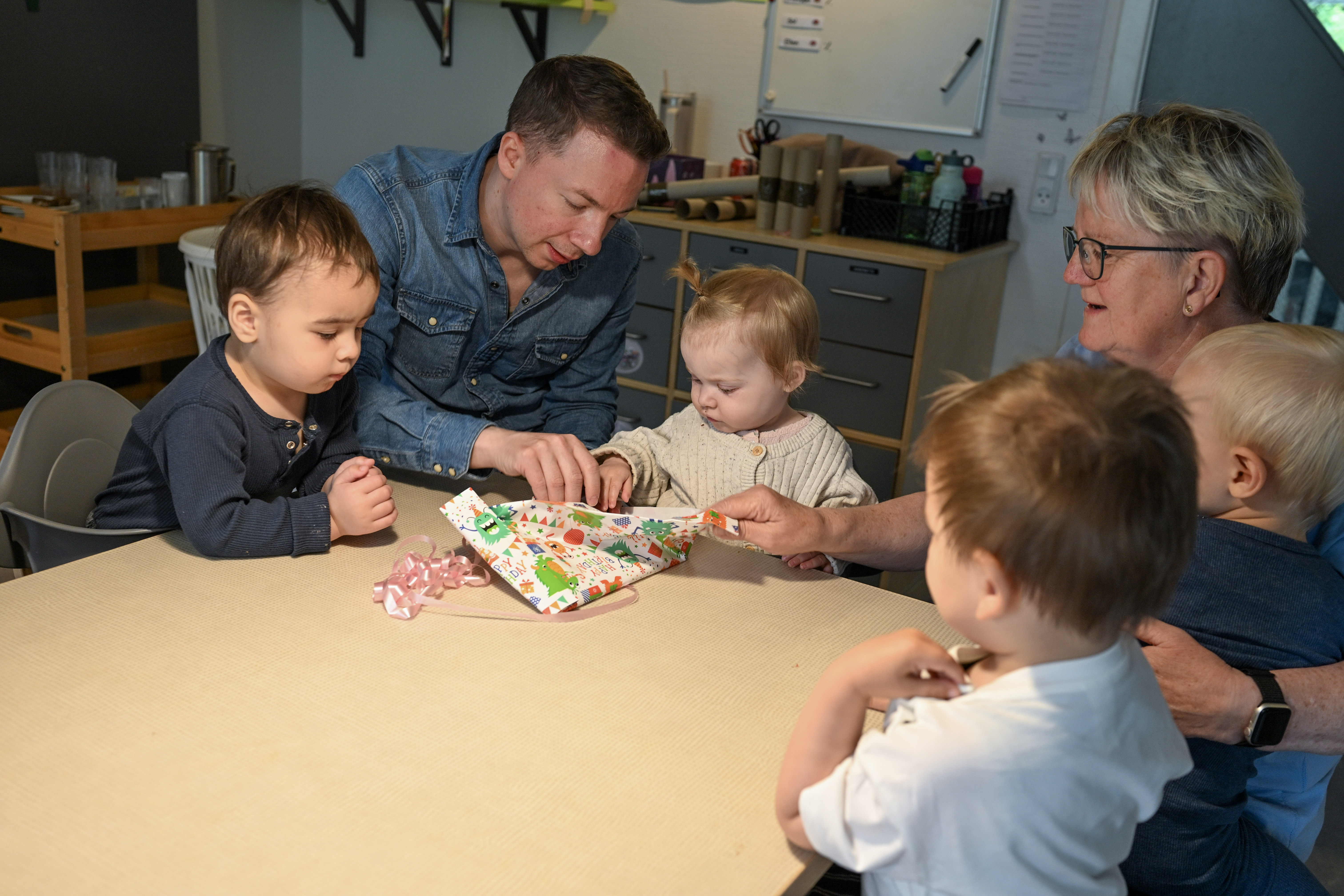 To voksne hjælper barn med at pakke en gave op, mens andre børn kigger på