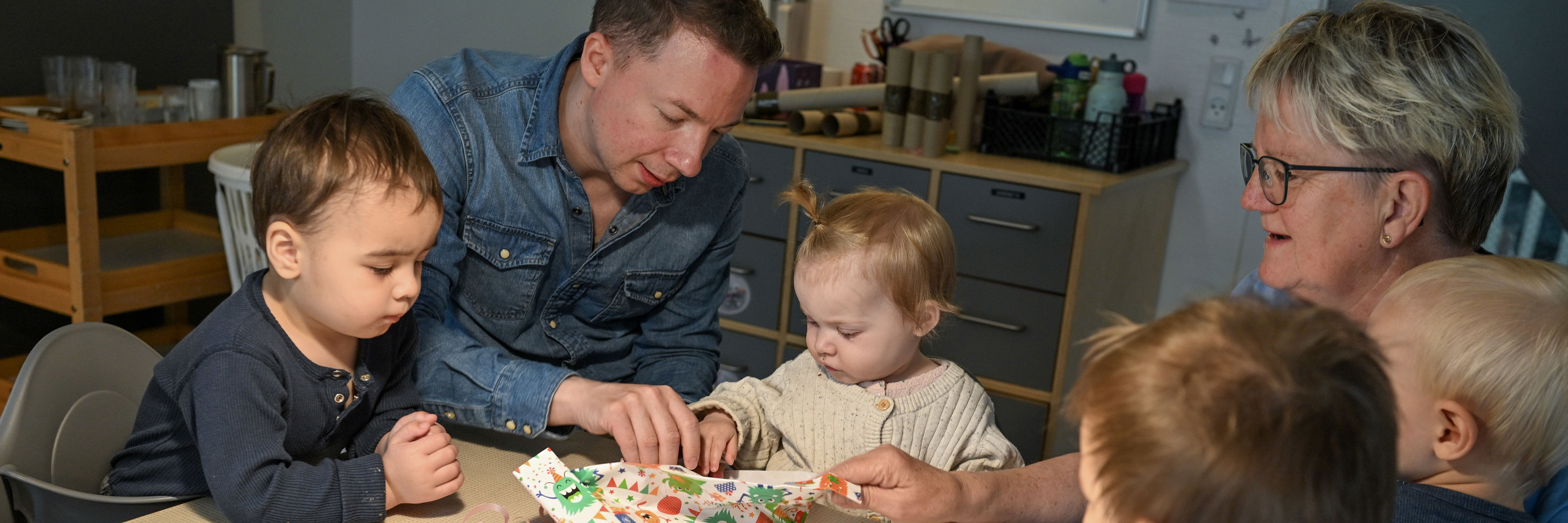 To voksne hjælper barn med at pakke en gave op, mens andre børn kigger på